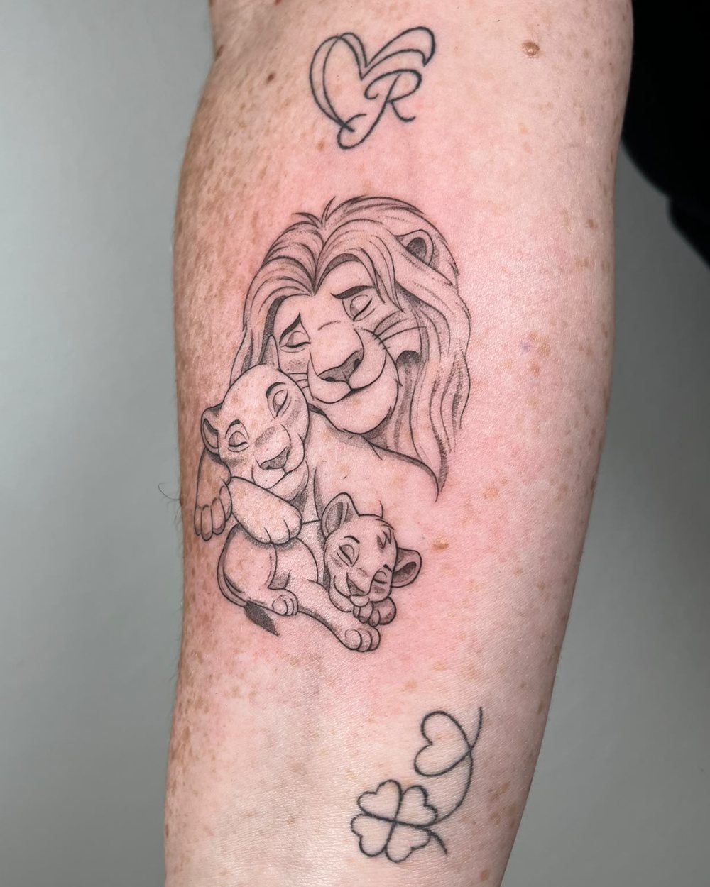 Tatuaje de línea fina del Rey León con sus cachorros, un corazón con una R y un trébol en el antebrazo